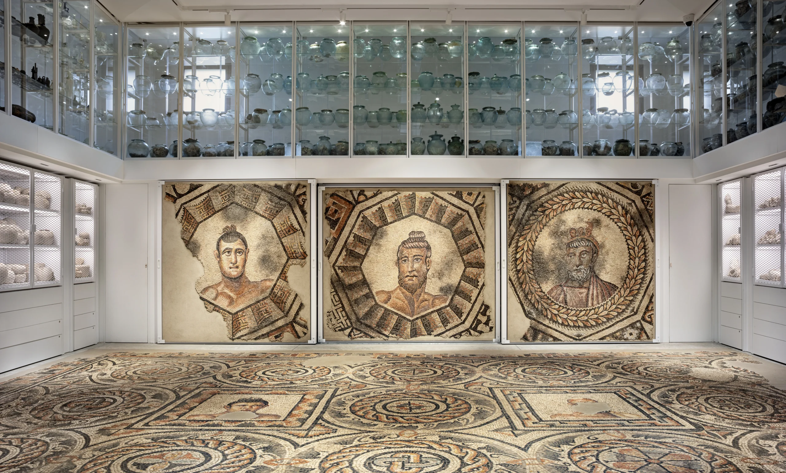 8031M_AC_2505 Veduta della sala centrale dei depositi del museo con mosaici a terra e alle pareti raffiguranti motivi geometrici e busti di atleti e visione delle vetrine del piano soppalcato con olle in vetro.